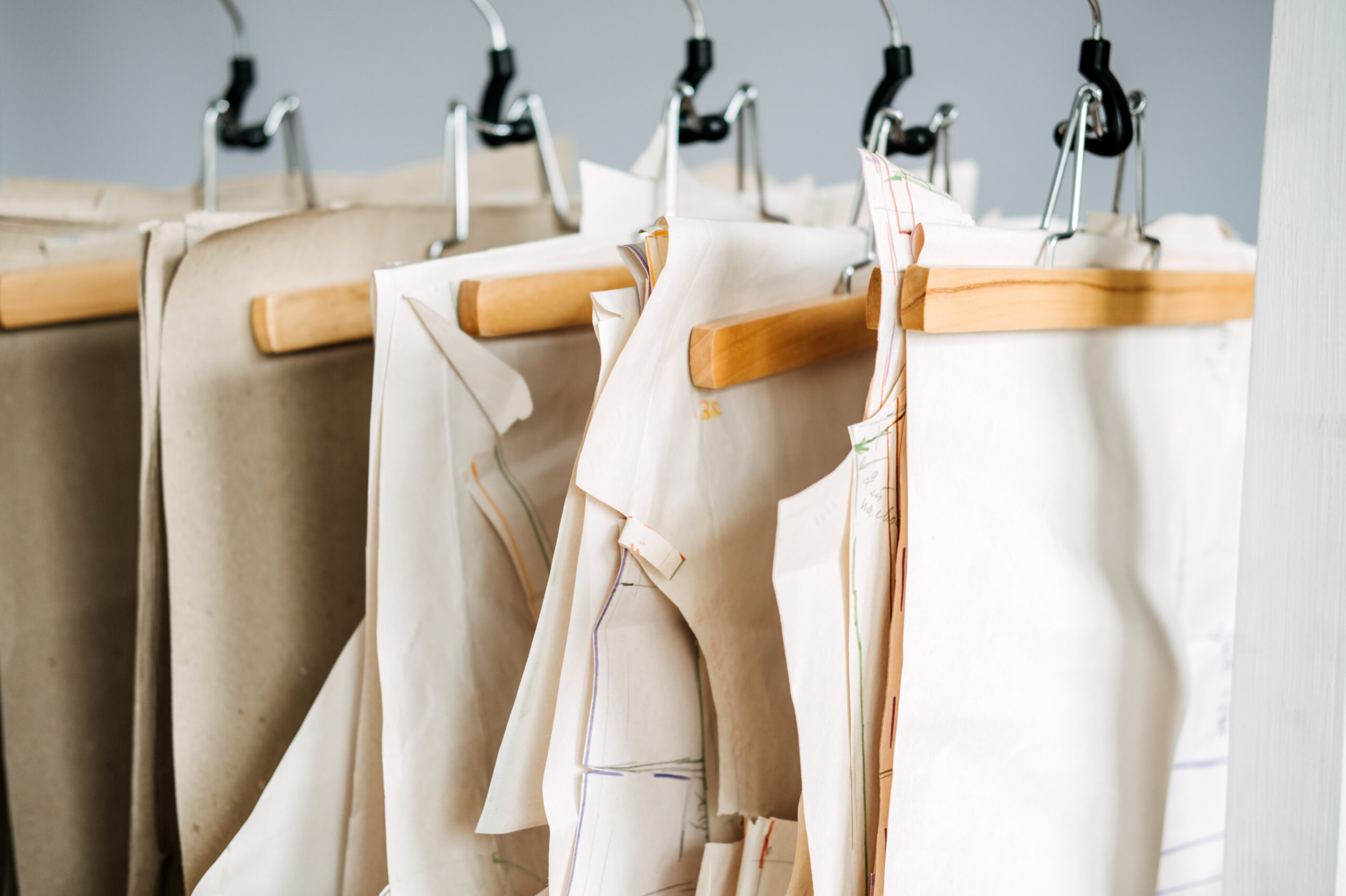 Many paper sewing patterns for different clothes hanging on the rack in sewing factory background. Clothing pattern, manufacture on sewing factory. Tailoring, small business.